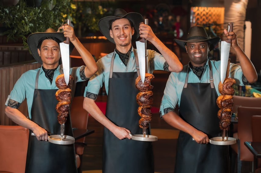 Three smiling servers holding large meat skewers