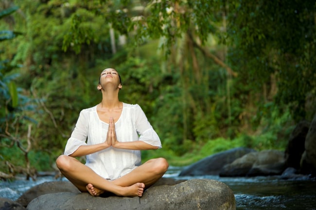 一名身穿白色无袖上衣的女士盘腿坐在雨林间的一块巨石上冥想