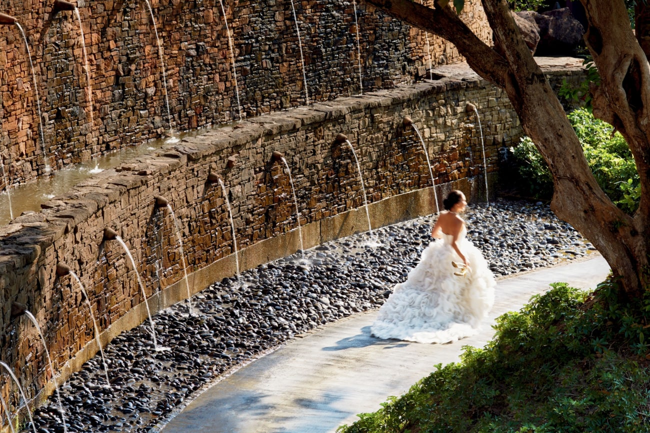 Bride in a strapless white gown with a full skirt glides by a stacked stone wall with water spouts