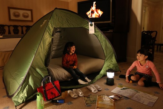 Two children play in and near a green tent set up in a suite with amenities like slippers, coloring books and a lantern
