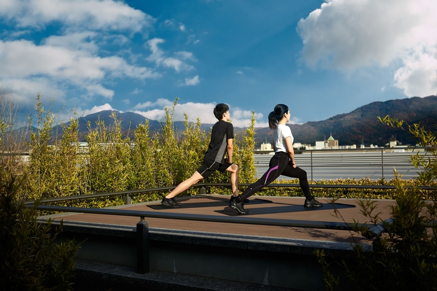 A man and woman in black workout clothes stretch on an open-air platform overlooking the mountains