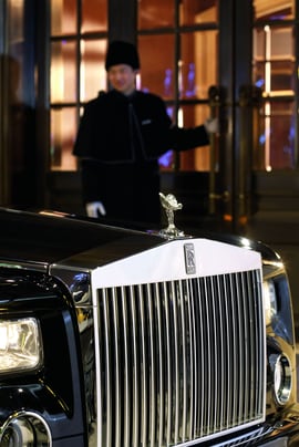 A luxury car pulls up to the front of a building where a doorman holds the door